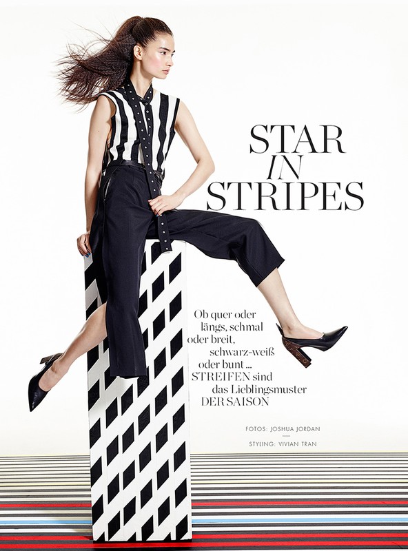 Fashion editorial shot of an Asian woman posing in a striped shirt and black pants, balanced on a striped block in a bright, white photo studio.
