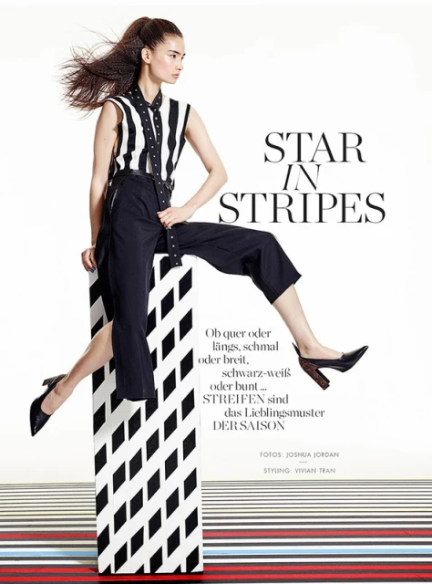 Fashion editorial shot of an Asian woman posing in a striped shirt and black pants, balanced on a striped block in a bright, white photo studio.