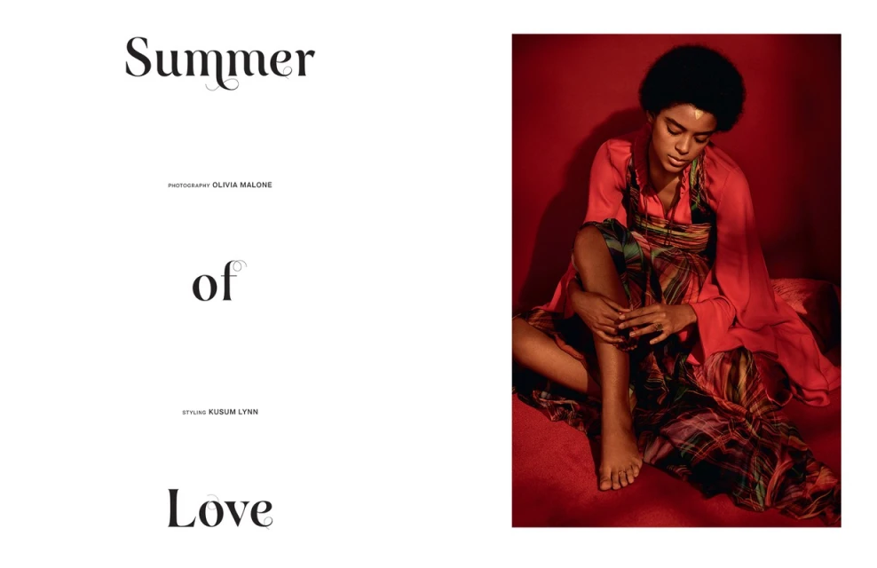 A dark-skinned model with an afro poses indoors in a patterned dress with a red top, sitting against a vibrant red wall. Fashion.