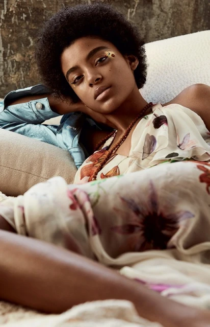 Portrait of a serene young woman with an afro and floral dress, reclining on a cream couch with a textured wall in the background.