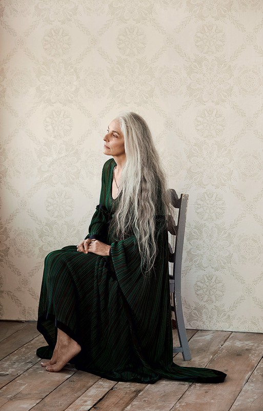 Elderly woman with long grey hair wearing a dark green pleated dress seated on a grey wooden chair in a room.
