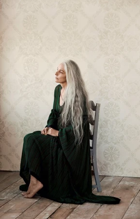 Elderly woman with long grey hair wearing a dark green pleated dress seated on a grey wooden chair in a room.