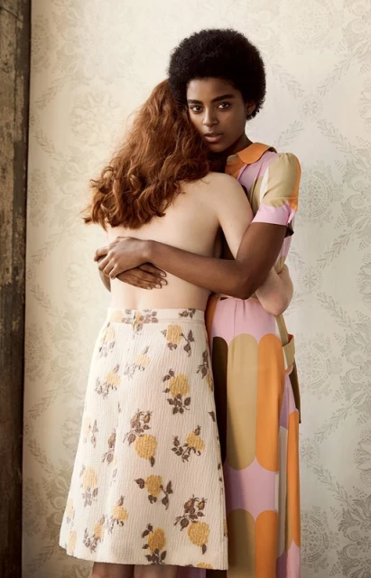 Two women embrace indoors against patterned wallpaper. One woman, facing forward, has dark skin and short hair and wears a patterned dress.