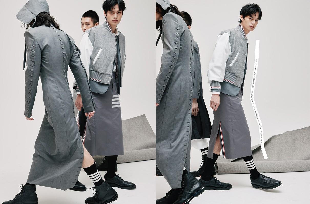 Three male models showcasing fashion designs featuring a long grey overcoat, a skirt, black socks with white stripes and black boots.