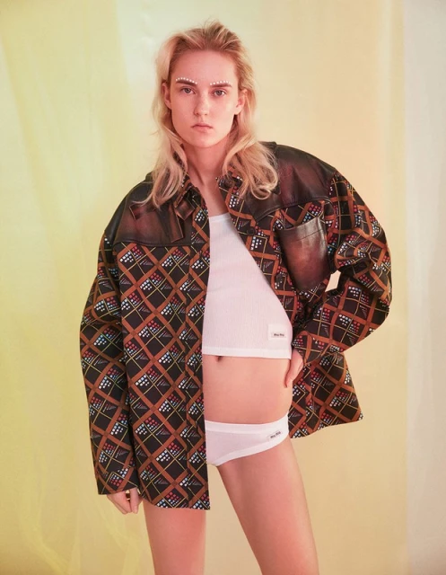 A blonde woman wears a patterned shirt and white undergarments while posing indoors against a background with soft yellow and white lighting.