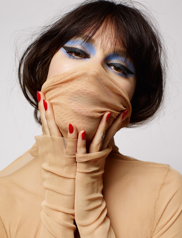 Portrait of a woman with dark hair and blue eyeshadow posing with a tan, sheer fabric covering her face and hands in a studio shot.