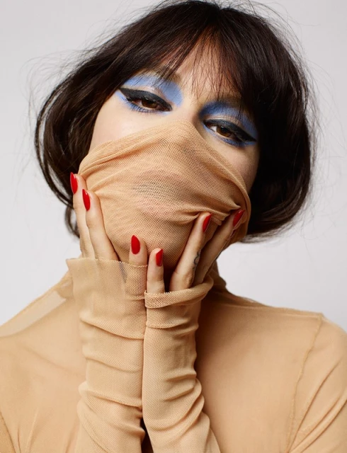 Portrait of a woman with dark hair and blue eyeshadow posing with a tan, sheer fabric covering her face and hands in a studio shot.