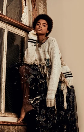 Black woman with short afro hair wearing a cream knit sweater and dark sheer skirt posed near an old wooden window frame.