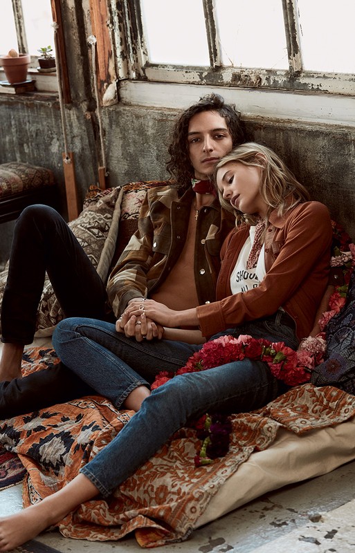 A young adult couple sits together on a rug in a room with sunlit windows. The man and woman wear casual 70s-inspired fashions.