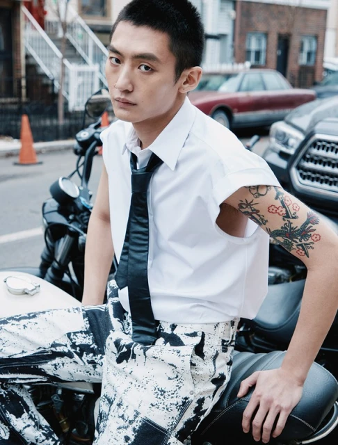Young adult Asian man posing on a motorcycle in an urban setting, wearing a white shirt and black and white patterned pants.