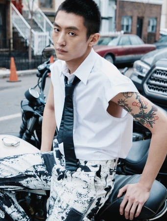 Young adult Asian man posing on a motorcycle in an urban setting, wearing a white shirt and black and white patterned pants.