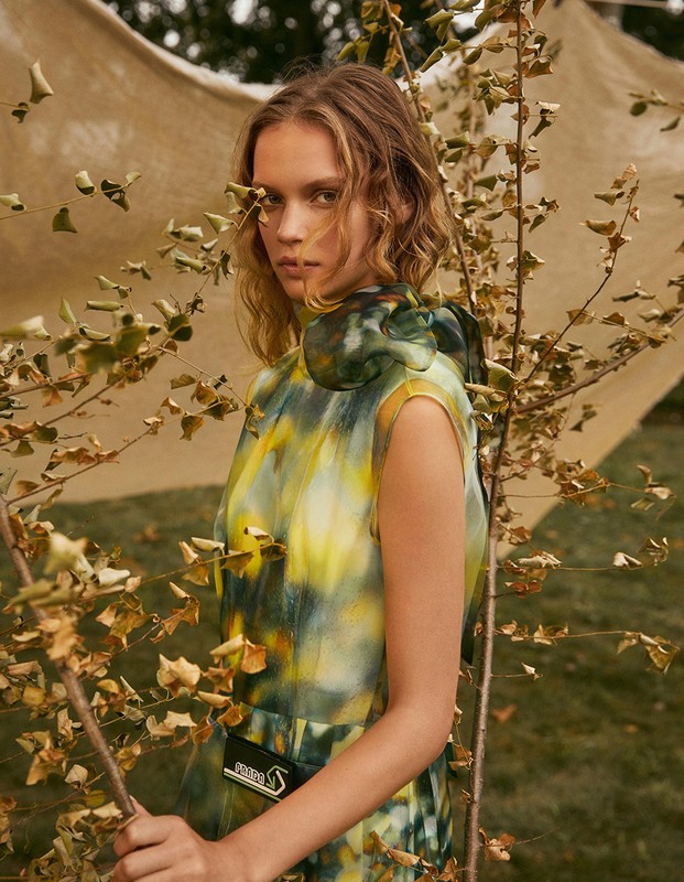 A fashionable woman in a yellow, white, and green dress poses outdoors among small trees and foliage, looking directly at the camera.