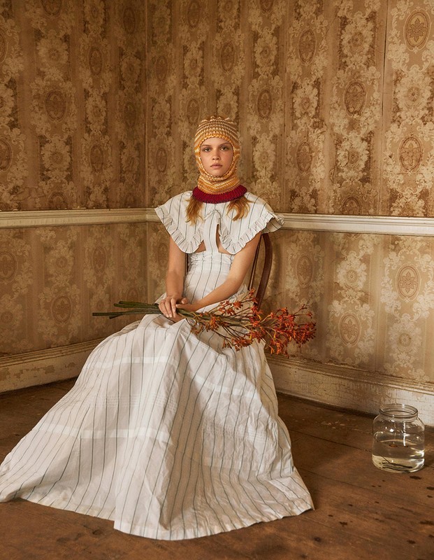 An adult female model is sitting in a striped dress, holding dried flowers against an old floral wallpaper backdrop and wood floor.
