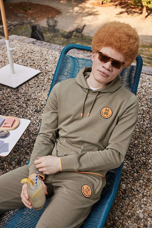 Man in olive tracksuit relaxing in blue deck chair outdoors, holding drink with lemon and straw, wearing sunglasses in dappled sunlight.