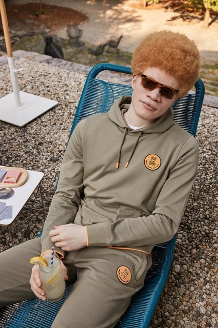 Man in olive tracksuit relaxing in blue deck chair outdoors, holding drink with lemon and straw, wearing sunglasses in dappled sunlight.