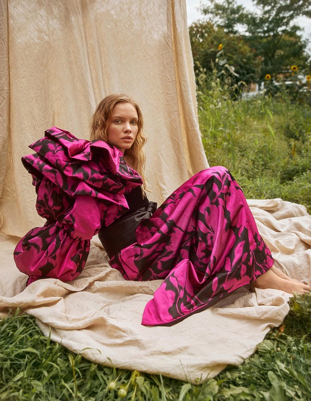 Young woman in a striking pink and black gown sits on a blanket in a grassy field, posing with a serious expression for an outdoor fashion editorial.