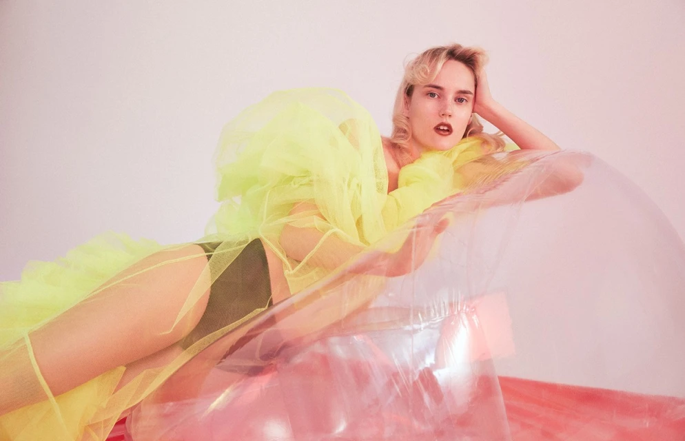 Blonde woman with open mouth reclines in an inflatable chair wearing a bright yellow tulle wrap against a pale pink studio backdrop.