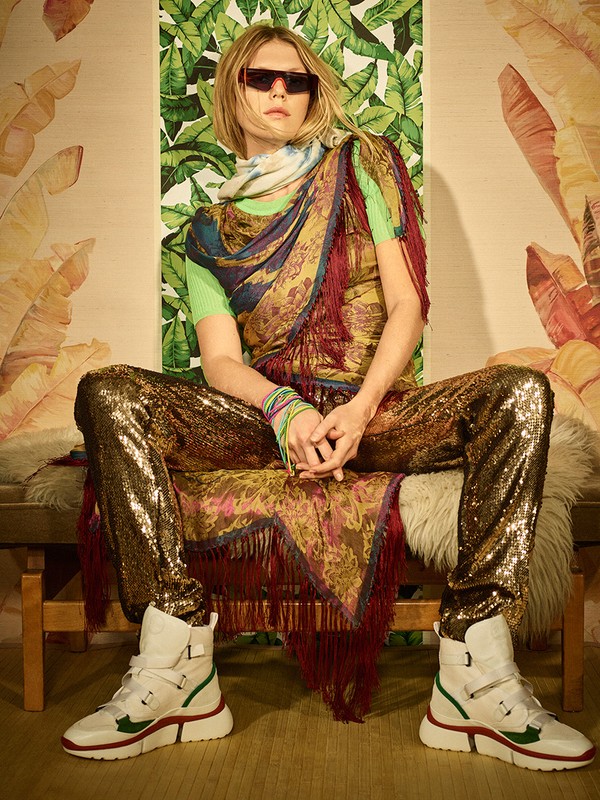 A person in sunglasses, sequined pants, and high-top sneakers is sitting on a wooden bench in front of a leafy wallpaper backdrop.