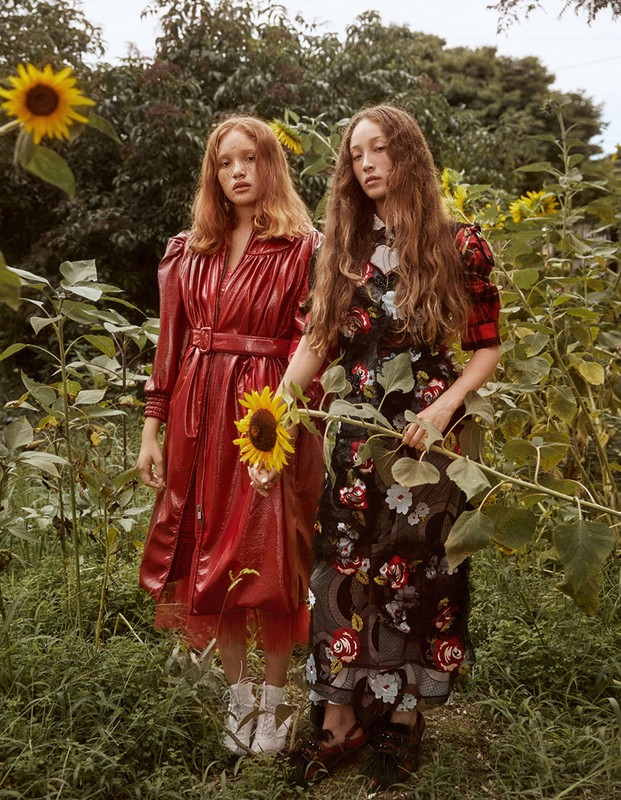 Two women in floral and red outfits pose in a field of sunflowers, exuding a fashion editorial vibe in an outdoor setting.