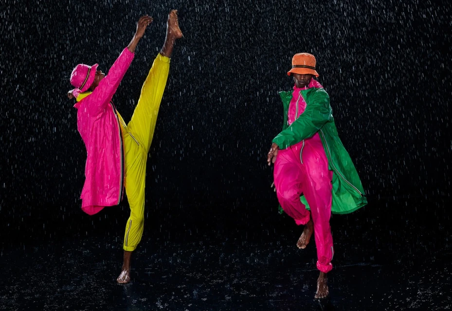 Two barefoot adult models in a downpour wear bright outerwear and bucket hats; one leaps while the other poses in rain.