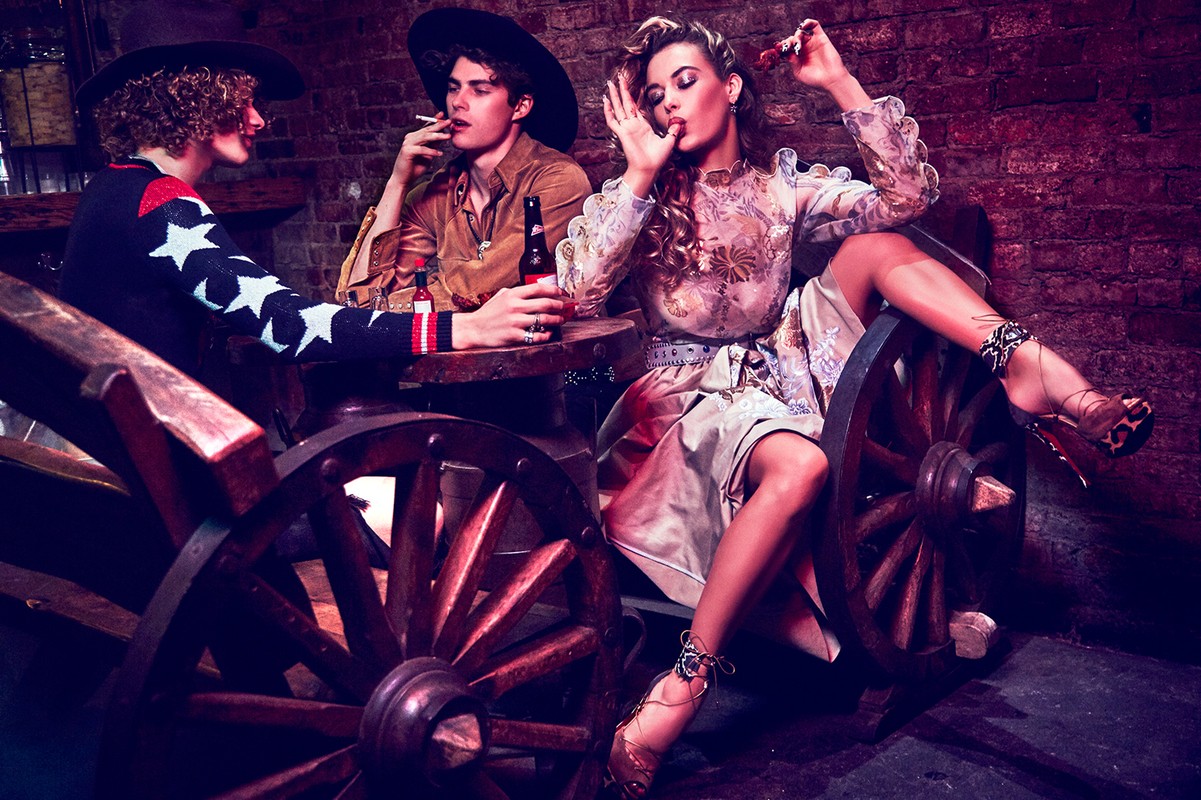 Three people, two men and a woman, sit around a small table. The woman is wearing a floral dress and heels while the men are smoking.