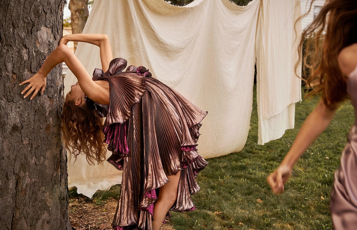 A woman in a metallic, ruffled dress poses next to a tree while another woman walks in the background, captured outdoors with soft light.