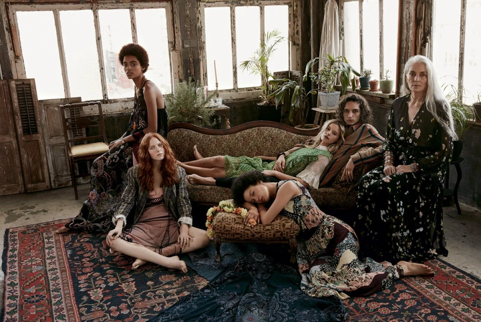 Six women of diverse ethnicities and ages in floral print dresses are posing on a patterned rug and vintage sofa indoors in natural light.