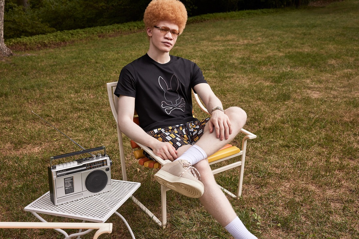Albino man with afro sits outdoors on a chair in a graphic tee and patterned shorts with a vintage boombox radio on a table.