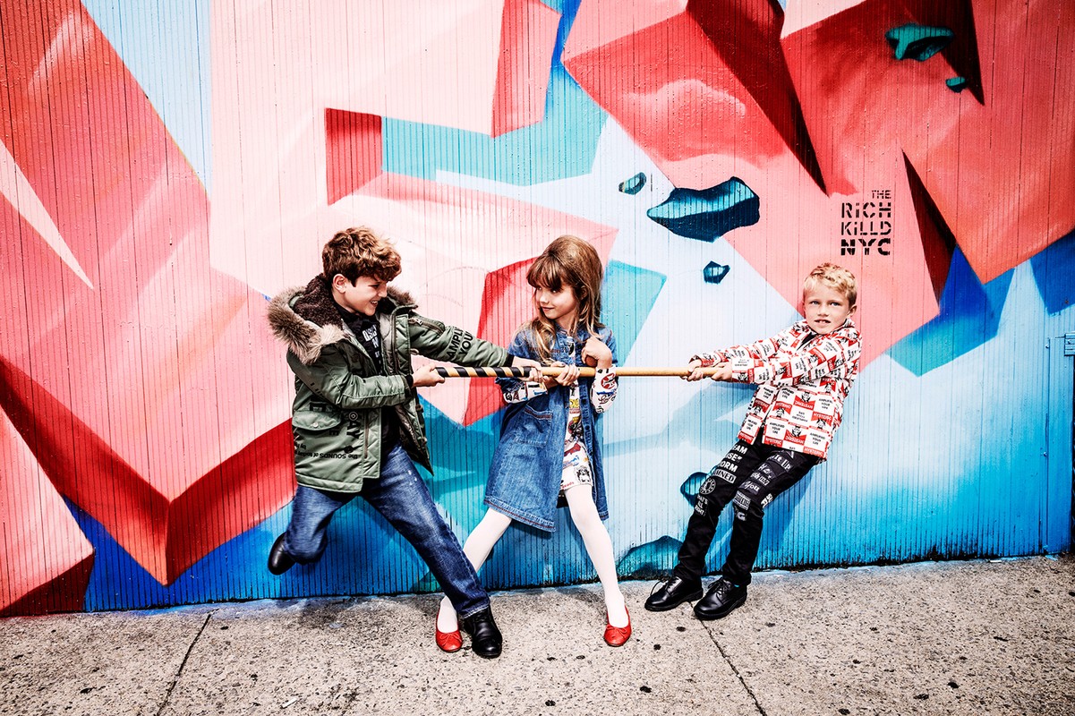 Three children playfully engage in a tug-of-war against a colorful graffiti wall, all clad in stylish clothing and red shoes.