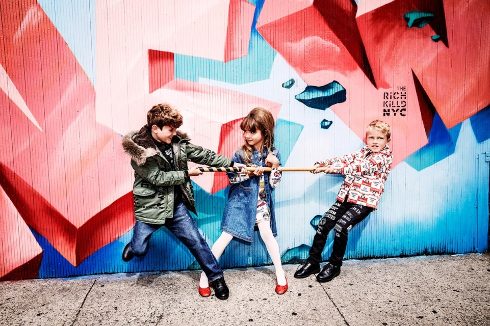 Three children playfully engage in a tug-of-war against a colorful graffiti wall, all clad in stylish clothing and red shoes.