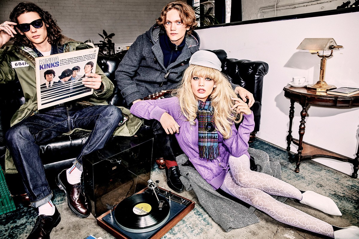 Three fashion models pose in an indoor setting, one holds a vinyl record cover, next to a vintage record player and a decorative table lamp.