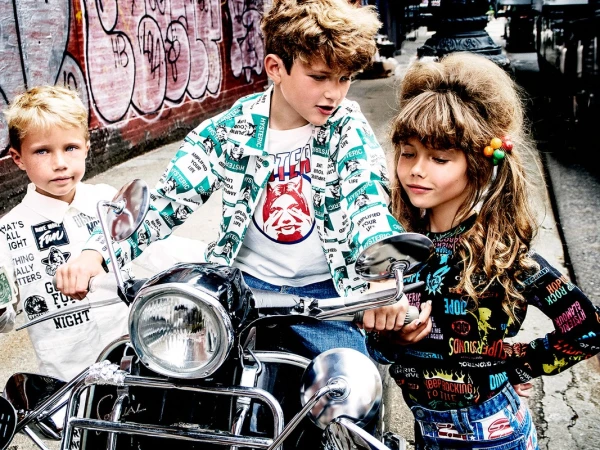 Three children posing with a dark motor scooter in an urban alleyway featuring graffiti art on brick walls.