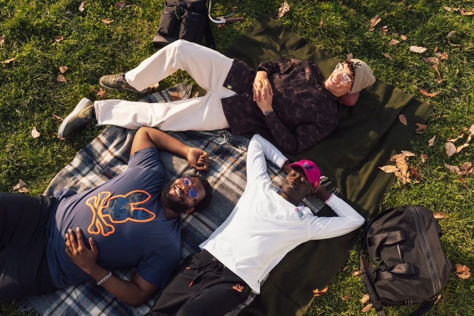 Three men lie on a plaid blanket in a grassy park, wearing casual clothing, and sunglasses on a sunny day, relaxed and enjoying the outdoors.
