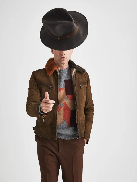Adult male in a brown shearling jacket, brown pants, and a gray geometric patterned sweater pointing directly at the camera with a hat on his head.