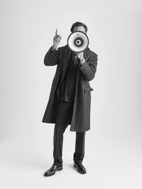 Full studio shot of a man in a striped suit and plaid overcoat holding a megaphone to his face, pointing up against a white backdrop.