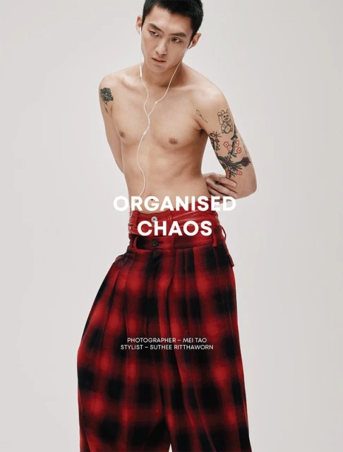 Shirtless man in studio with tartan pants, listening to earbuds, against a white backdrop with graphic designer and stylist credit.