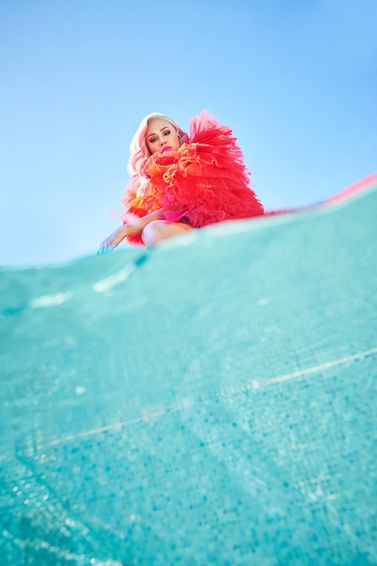 Woman with pink hair and red tulle dress posing in a squat on a blue and white patterned surface against a light blue sky.