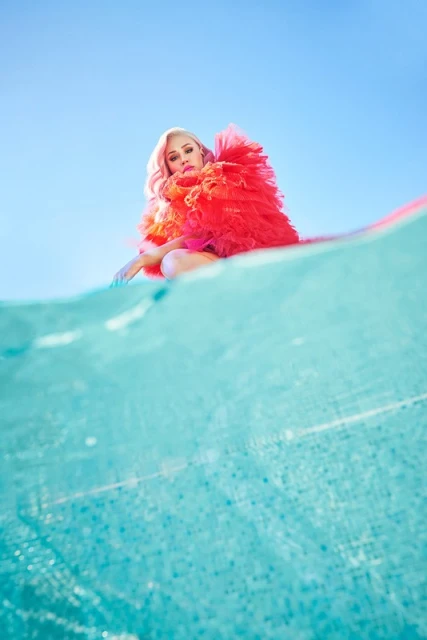 Woman with pink hair and red tulle dress posing in a squat on a blue and white patterned surface against a light blue sky.