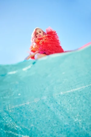 Woman with pink hair and red tulle dress posing in a squat on a blue and white patterned surface against a light blue sky.