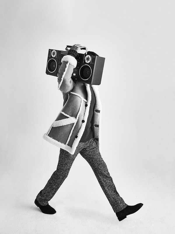 Person in coat walking with vintage boombox over face in black and white, in a bright, neutral studio, fashion concept.