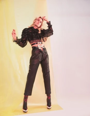 A striking fashion shot of a blonde adult woman in a sheer black top, black leather pants, and booties, against a yellow and white backdrop.
