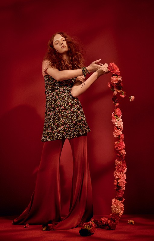 Young woman with long red hair poses with a cascade of red and pink carnations against a red background, wearing a floral dress and wide-leg pants.