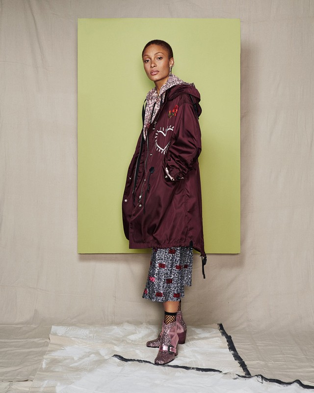 Woman in burgundy coat with heart design standing in a studio setting, wearing patterned dress and sequin boots in a studio shot.
