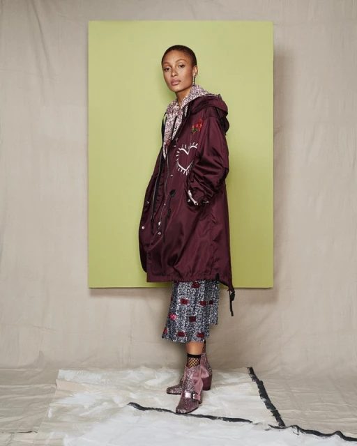 Woman in burgundy coat with heart design standing in a studio setting, wearing patterned dress and sequin boots in a studio shot.