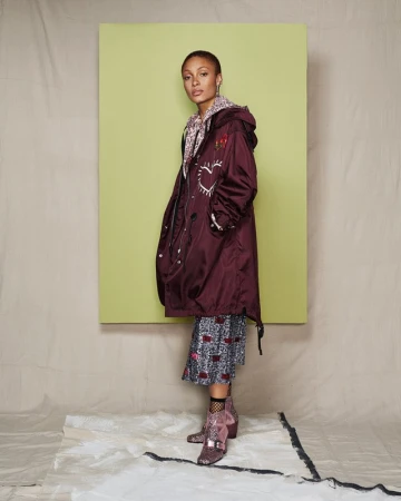 Woman in burgundy coat with heart design standing in a studio setting, wearing patterned dress and sequin boots in a studio shot.