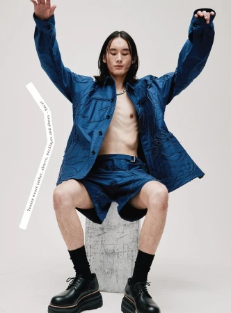 Model with long dark hair wearing blue jacket and shorts with black shoes seated against white background