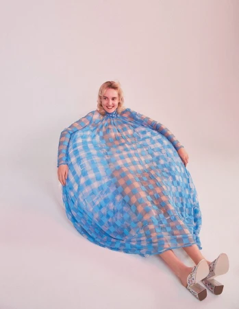 Model sitting in a voluminous blue and white checkered dress against a pale background with heeled mules