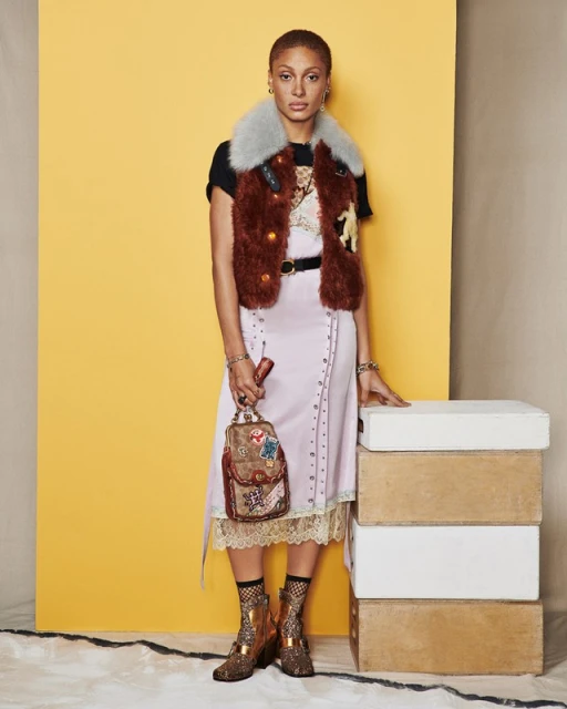 A young woman stands indoors against a colorful backdrop. She is wearing a fur vest, dress with lace trim, patterned socks, and platform shoes.