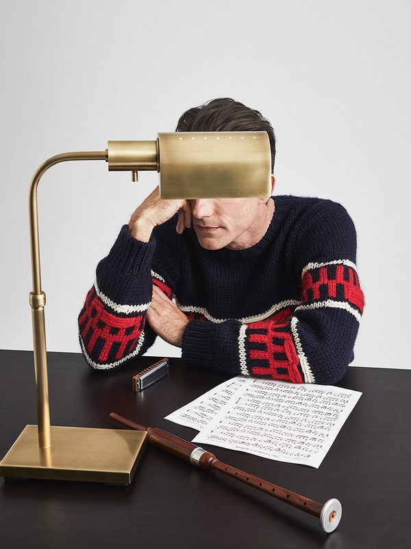 Man in a patterned sweater sits at a desk with a lamp, sheet music, a bagpipe, and a harmonica. Indoor lighting. Tabletop setting.