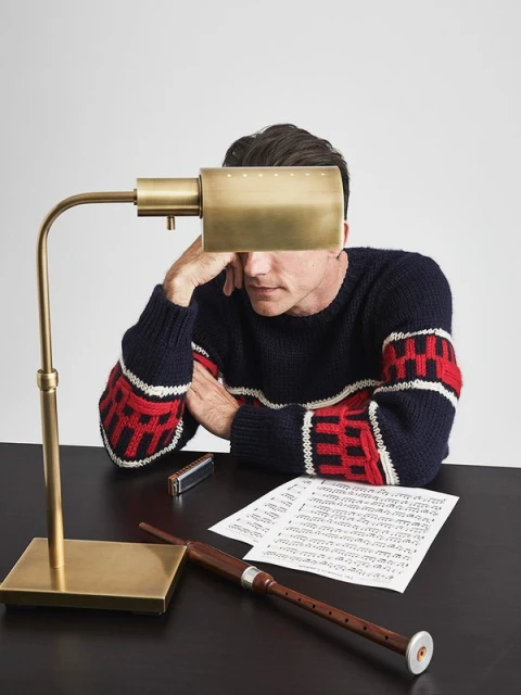 Man in a patterned sweater sits at a desk with a lamp, sheet music, a bagpipe, and a harmonica. Indoor lighting. Tabletop setting.